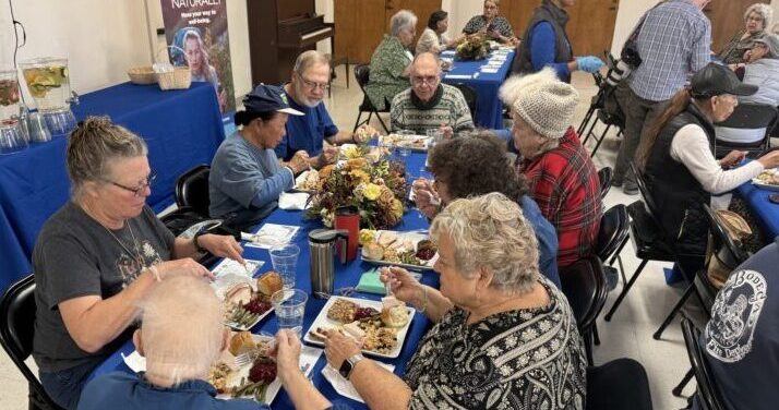 Older adults sitting at a long table with a blue tablecloth, eating a thanksgiving meal.